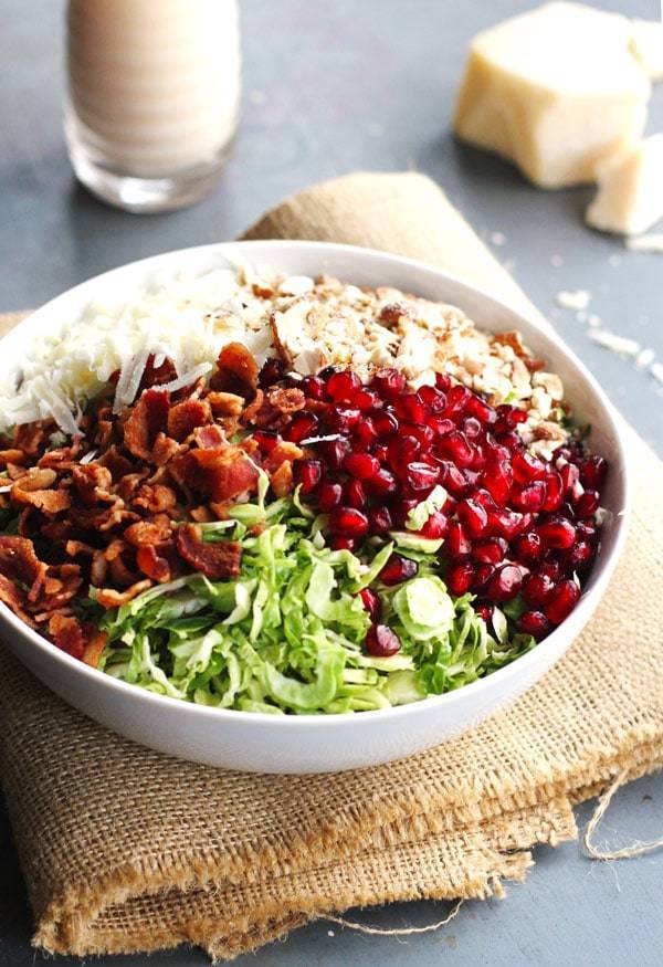 Brussels sprout salad has pomegranates, almonds, crumbled bacon, and homemade creamy salad dressing in a white bowl.