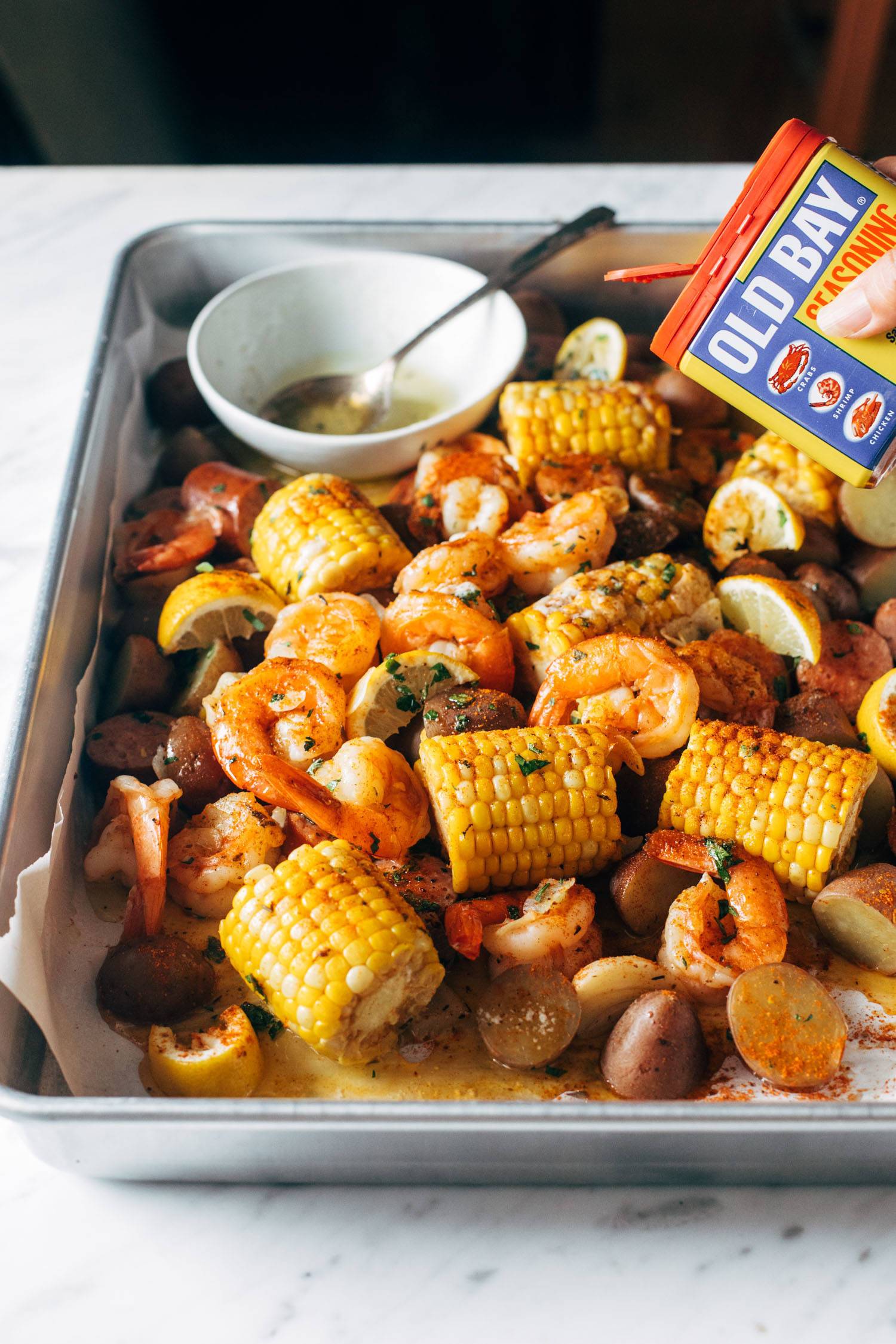 A shrimp boil on a sheet pan with garlic infused butter. A white hand is sprinkling Old Bay seasoning on top.